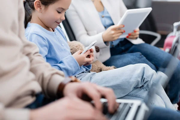 Cheerful girl using smartphone near blurred parents with gadgets — Fotografia de Stock