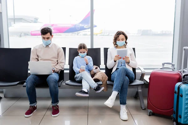 Family in medical masks using gadgets in airport — Stock Photo
