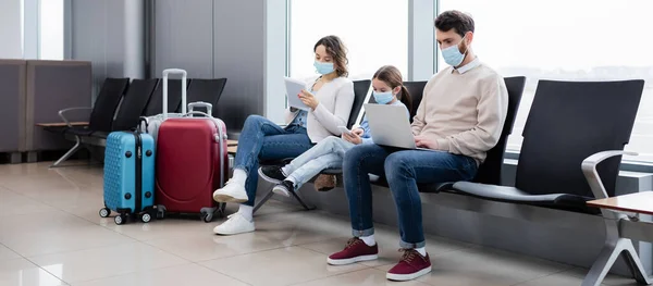 Family in medical masks using gadgets in airport, banner — Stock Photo