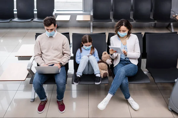 Kind und Eltern in medizinischen Masken mit Gadgets am Flughafen — Stockfoto