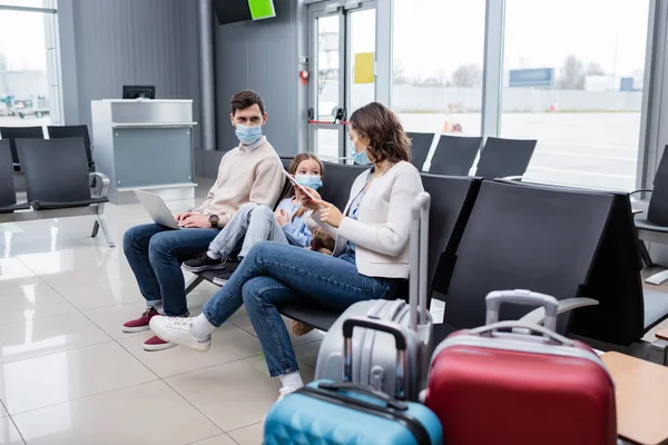 Niño y padre en máscaras médicas sosteniendo gadgets y mirando a la mujer en el aeropuerto - foto de stock