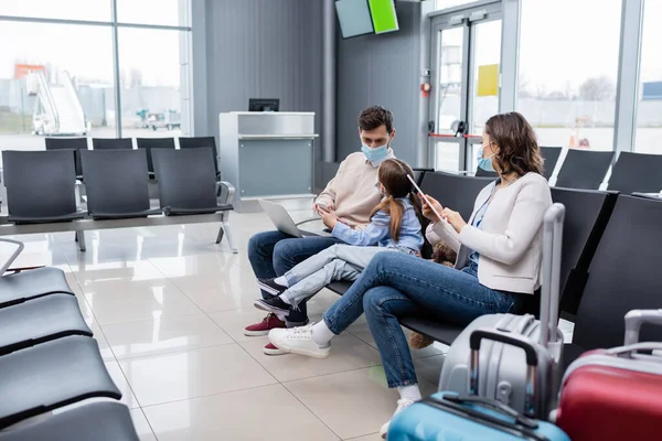 Kid in medical mask showing smartphone to father in airport — стоковое фото