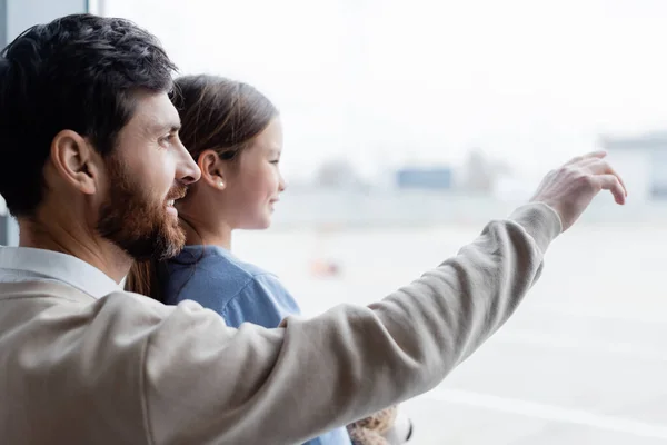 Happy father pointing at window near smiling daughter in airport — стоковое фото