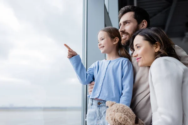 Happy kid pointing with finger at window near cheerful parents in airport — стоковое фото