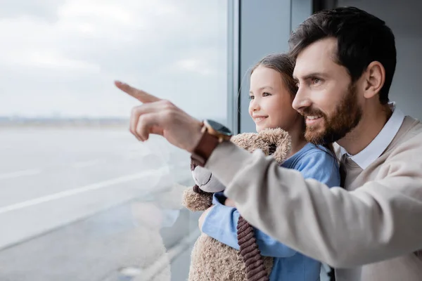 Glücklicher Mann zeigt mit Finger auf Fenster nahe Tochter mit Plüschtier am Flughafen — Stockfoto