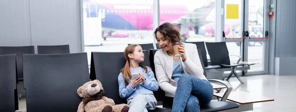 Kind hält Smartphone in der Hand und blickt fröhliche Mutter mit Pappbecher in der Lounge des Flughafens an, Banner — Stockfoto