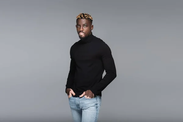 Trendy african american man with vitiligo skin standing with hands in pockets of jeans isolated on grey — Stock Photo