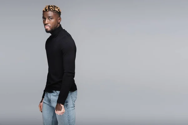 African american man with vitiligo and trendy hairstyle posing in jeans and black turtleneck isolated on grey — Stock Photo