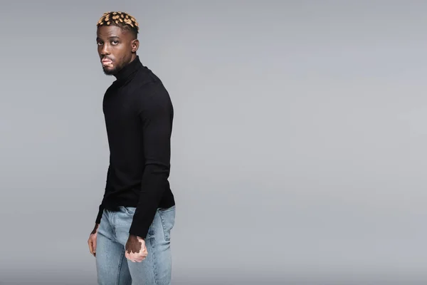 African american man with vitiligo and trendy hairstyle posing in jeans and black turtleneck isolated on grey — стоковое фото