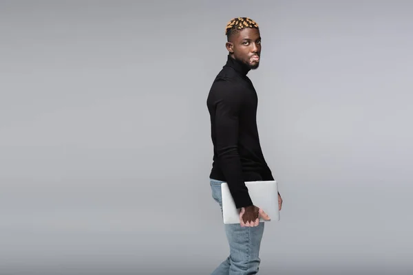 African american man with vitiligo holding laptop and looking at camera isolated on grey — Photo de stock