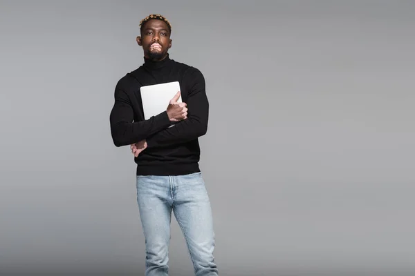 African american man with vitiligo, wearing black turtleneck and jeans, holding laptop isolated on grey — стоковое фото