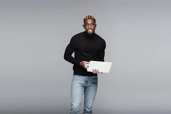 Афроамериканський чоловік з вітіліго тримає ноутбук, стоячи в джинсах і чорному черепашку ізольовано на сірому — стокове фото