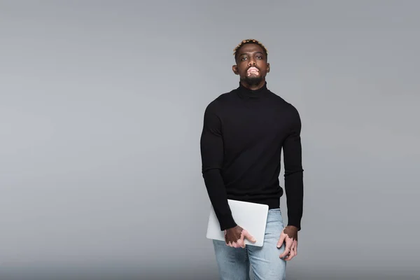 Stylish african american man with vitiligo looking at camera while holding laptop isolated on grey — Stock Photo