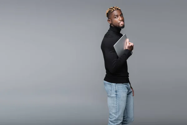 African american man with vitiligo, wearing jeans and black turtleneck, holding laptop isolated on grey — Stockfoto