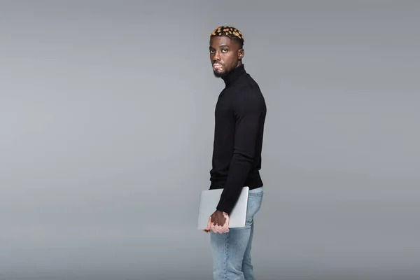Модний афроамериканський чоловік з вітіліго стоїть з ноутбуком і дивиться на камеру ізольовано на сірому — стокове фото
