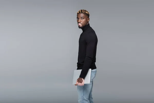 Trendy african american man with vitiligo standing with laptop and looking at camera isolated on grey — Stock Photo