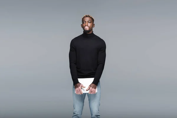 Joven afroamericano con vitiligo, en jeans y cuello alto negro, de pie con portátil aislado en gris - foto de stock