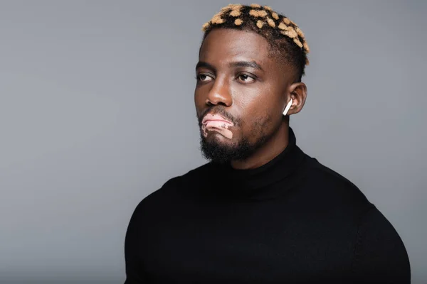 Stylish african american man with vitiligo looking away while listening music in earphone isolated on grey - foto de stock