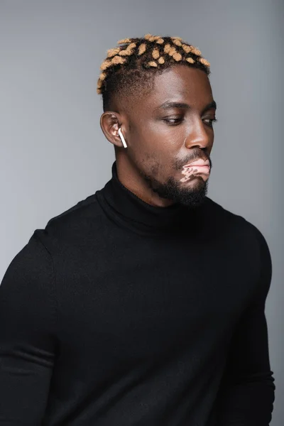 Young african american man with vitiligo and trendy hairstyle listening music in earphone isolated on grey — стоковое фото