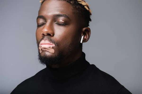 Young african american man with vitiligo and closed eyes listening music in earphone isolated on grey — Stockfoto