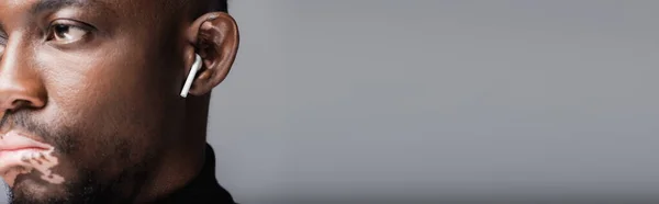 Cropped view of blurred african american man with vitiligo listening music in earphone isolated on grey, banner — Fotografia de Stock