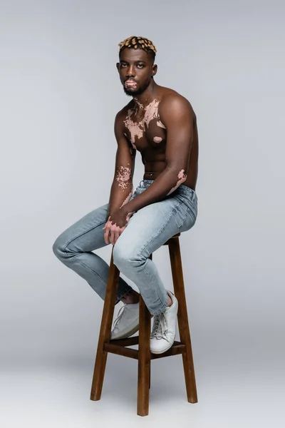 Shirtless african american man with vitiligo looking at camera while sitting on stool in jeans on grey — Foto stock