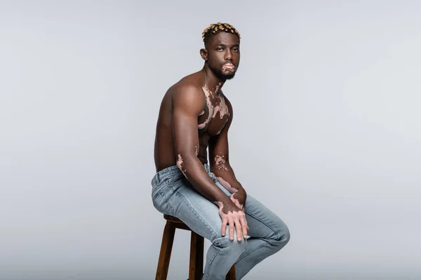 Без сорочки афроамериканський чоловік з вітіліго дивиться на камеру, сидячи на табуреті в джинсах ізольовані на сірому — стокове фото
