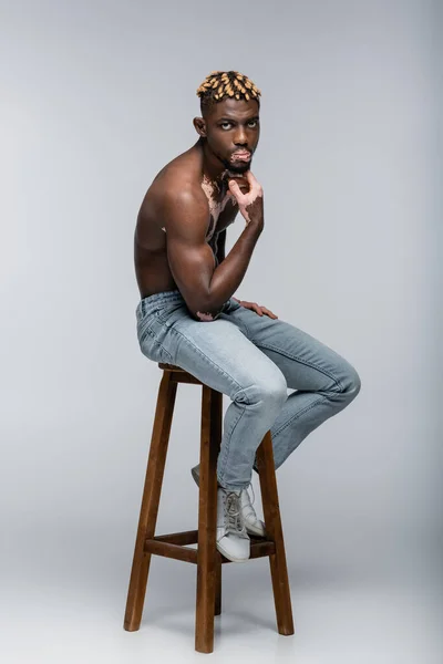 Hombre americano africano musculoso con piel de vitiligo cogida de la mano cerca de la barbilla mientras está sentado en el taburete en jeans en gris - foto de stock