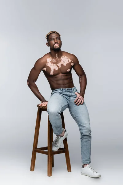 Shirtless african american man with vitiligo and stylish hairstyle posing on high stool on grey — Stock Photo