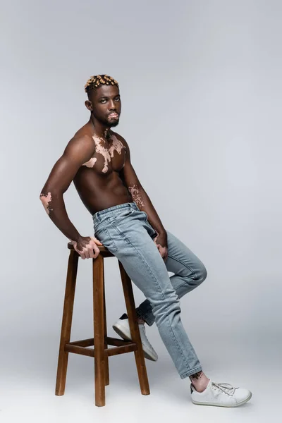 Афроамериканський чоловік з вітіліго позує без сорочки біля високого табурета на сірому — стокове фото