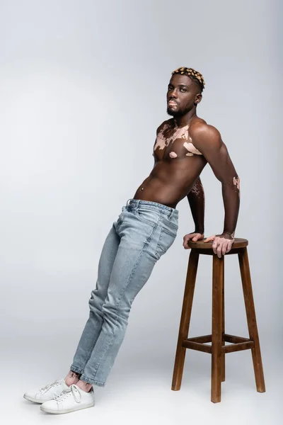 Hombre afroamericano sin camisa con vitiligo y torso muscular posando en jeans cerca de taburete alto en gris - foto de stock