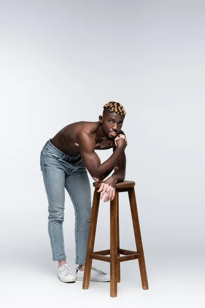 Shirtless african american man with vitiligo skin leaning on high stool while posing in jeans on grey — Photo de stock