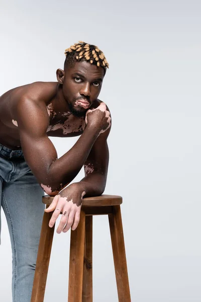Hombre americano africano sin camisa y musculoso con vitiligo posando con la mano cerca de la cara cerca de taburete de madera en gris - foto de stock