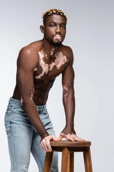 African american man with vitiligo skin and trendy hairstyle standing shirtless near wooden stool on grey — Foto stock
