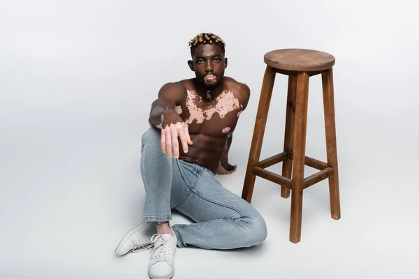 Hombre afroamericano con vitiligo y torso sin camisa sentado cerca de taburete alto en gris - foto de stock