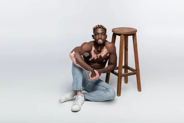М'язистий афроамериканський чоловік зі шкірою вітіліго і стильною зачіскою, що сидить без сорочки біля дерев'яного табурета на сірому — стокове фото