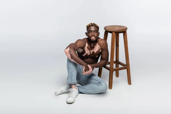 Muscular african american man with vitiligo skin and stylish hairstyle sitting shirtless near wooden stool on grey — стоковое фото