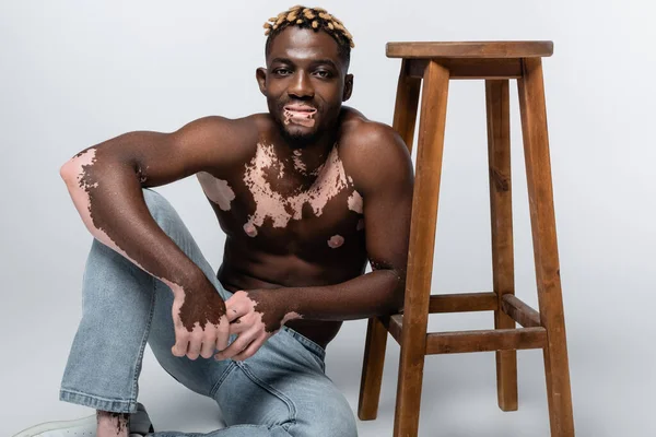 Усміхнений афроамериканський чоловік зі шкірою вітіліго та м'язистим торсом, що сидить біля дерев'яного табурета на сірому — стокове фото