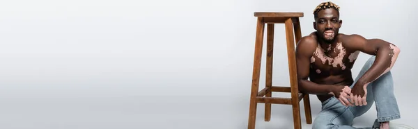 Smiling african american man with vitiligo skin sitting shirtless near wooden stool on grey, banner — Foto stock