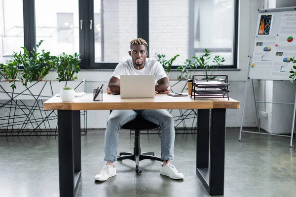 Joven afroamericano hombre con piel vitiligo sentado en el lugar de trabajo cerca de la computadora portátil y documentos en la oficina - foto de stock