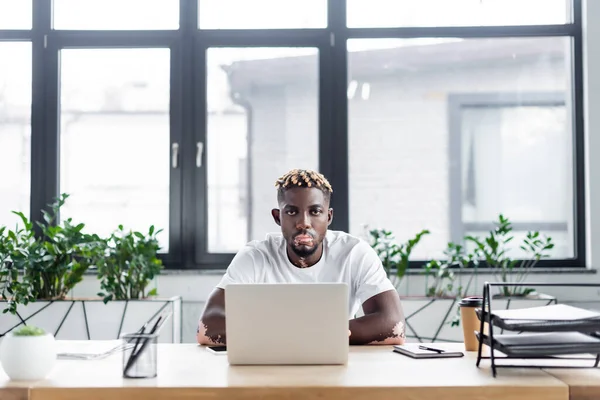 African american man with vitiligo and stylish hairstyle looking at camera near laptop and coffee to go in office — Foto stock