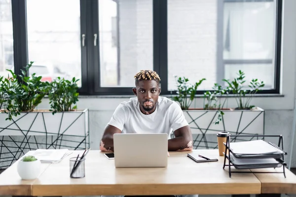 Hombre afroamericano con estilo vitiligo sentado en el lugar de trabajo cerca de la computadora portátil en la oficina - foto de stock