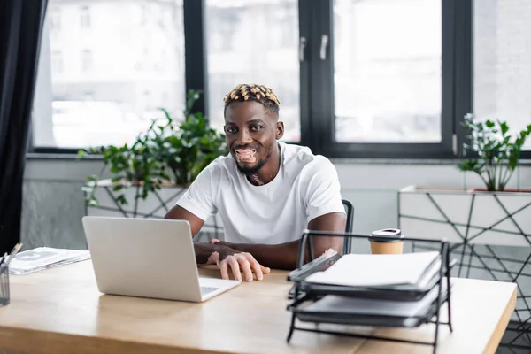 Hombre afroamericano con vitiligo sonriendo cerca del ordenador portátil, café para llevar y documentos en la oficina - foto de stock