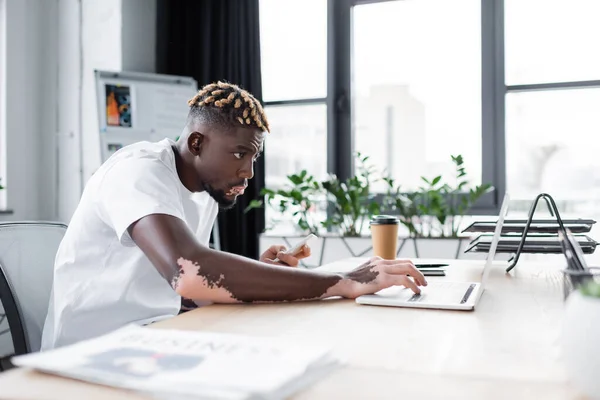 Hombre afroamericano concentrado con vitiligo celebración de teléfono inteligente mientras escribe en el ordenador portátil en la oficina - foto de stock