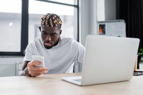 Joven afroamericano hombre con vitiligo mirando el teléfono móvil cerca borrosa portátil en la oficina - foto de stock