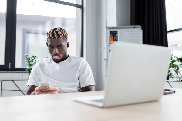 Молодий афроамериканський чоловік з вітіліго обміну повідомленнями на мобільному телефоні біля розмитого ноутбука в офісі — стокове фото