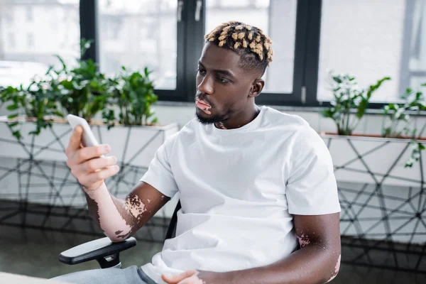 Молодий афроамериканський чоловік з вітіліго і модною зачіскою дивиться на мобільний телефон, сидячи в офісі — стокове фото