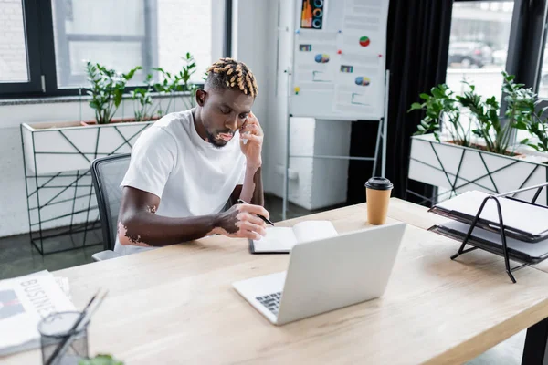 African american man with vitiligo skin talking on smartphone near notebook and laptop in office — Foto stock