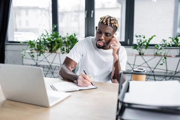 Hombre afroamericano con estilo vitiligo hablando en el teléfono inteligente y la escritura en el portátil cerca borrosa - foto de stock