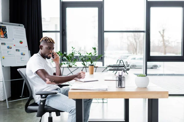 Афроамериканський чоловік з вітіліго розмовляє на мобільному телефоні біля ноутбука і фліп-чарт в офісі — стокове фото